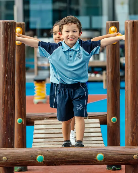 A young child stands at the top of a wooden climbing frame in the playground with both arms raised