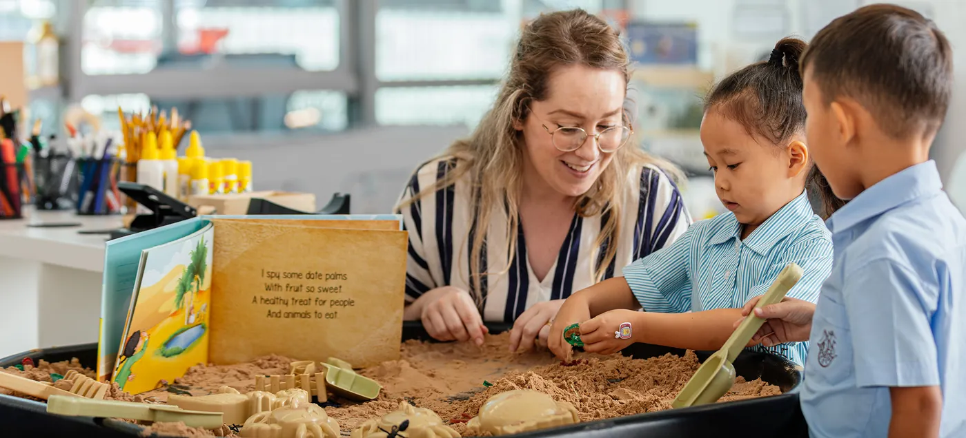 A teacher kneels at a low table reading with two young children in the Lower Junior School