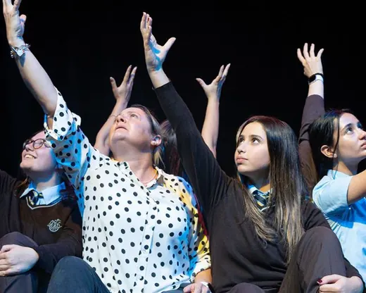 Senior students in black and white costume throw their arms upwards on stage during a drama production
