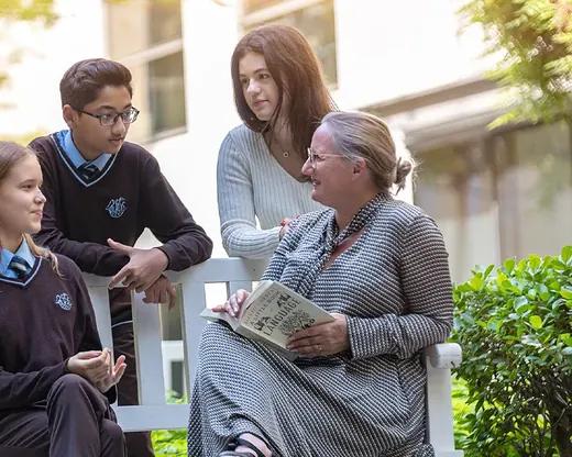A teacher sits talking with three senior students on a bench under a tree on campus